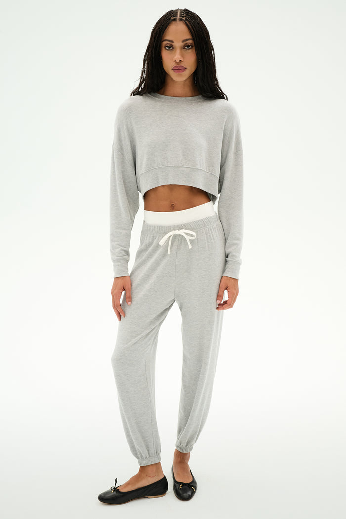 A woman stands against a plain background wearing SPLITS59's Andie Dual Waist Sweatpant in Heather Grey/White—soft modal blend joggers with a drawstring—paired with a light grey cropped sweatshirt and black ballet flats.