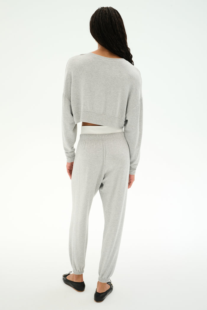 A person stands facing away, wearing SPLITS59’s Andie Dual Waist Sweatpant in Heather Grey/White with a matching light gray long-sleeve top and black flats, all crafted from a soft modal blend with spandex, against a white background.