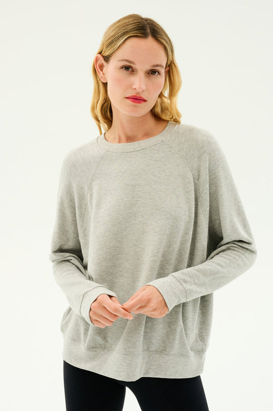 A person with blonde hair wearing a light gray Andie Fleece Sweatshirt - Heather Grey by SPLITS59 and black pants stands against a plain white background.
