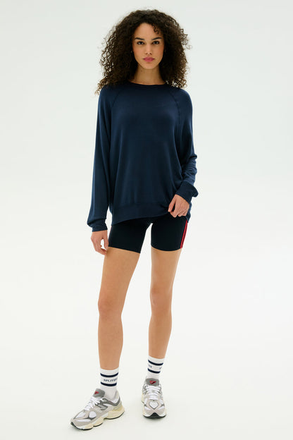 A woman faces forward in a SPLITS59 Andie Fleece Sweatshirt - Indigo, black shorts with a red stripe, white athletic socks, and gray sneakers against a plain light background.