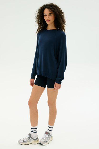 A person stands against a plain background wearing the SPLITS59 Andie Fleece Sweatshirt in Indigo, paired with black shorts, white socks, and silver sneakers.