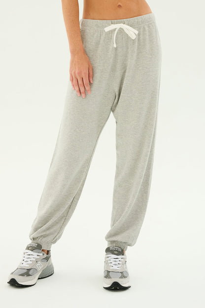 A person wearing the Andie Oversized Fleece Sweatpant in Heather Grey by SPLITS59 and gray sneakers stands on a plain background. The sweatpants, perfect for cool weather workouts or relaxing at home, feature an elastic waistband with a white drawstring. Only the lower half of the body is visible.