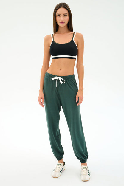 A woman faces forward in a black sports bra, SPLITS59's Andie Oversized Fleece Sweatpant in Military, and white sneakers—ideal for cool weather workouts—set against a plain white background.