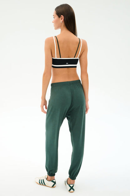 A woman faces away, wearing a black and white sports bra and SPLITS59 Andie Oversized Fleece Sweatpant in Military, paired with white sneakers with green stripes—ideal for cool weather workouts—on a plain light background.