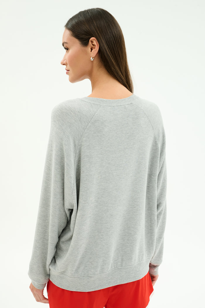 A woman stands with her back to the camera wearing the SPLITS59 Andie Oversized Fleece Varsity Graphic Sweatshirt in Heather Grey, paired with red shorts, set against a plain light background.
