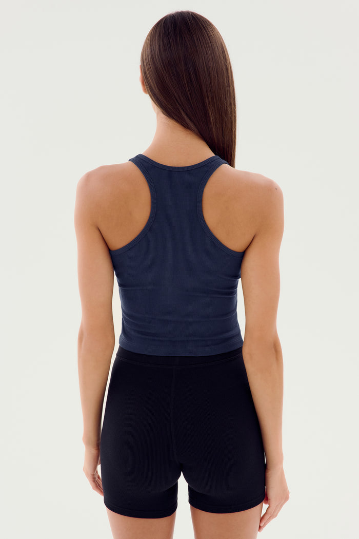 A person with long hair is standing with their back to the camera. They are wearing a dark blue SPLITS59 Ashby Rib Crop - Indigo and black shorts, perfect for gym workouts.