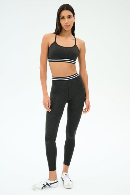 A woman poses in SPLITS59’s Bailey Active Rib High Waist 7/8 set in Heather Graphite, featuring a matching ribbed sports bra and leggings with white stripe accents, paired with white sneakers—ideal for gym workouts.