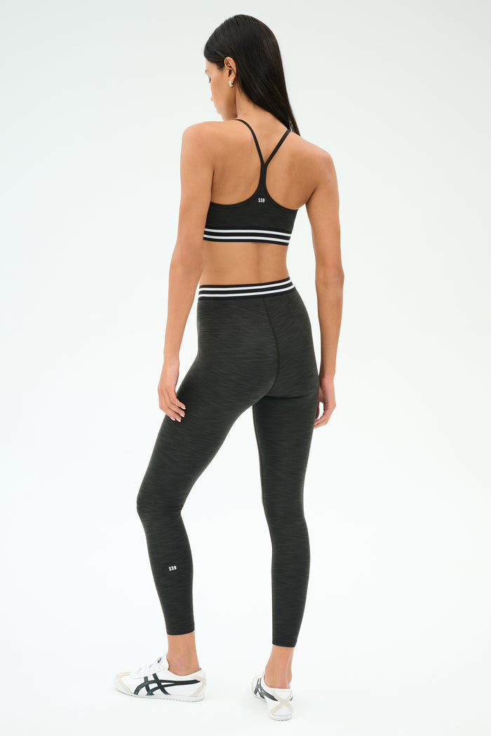 A woman wears the SPLITS59 Bailey Active Rib High Waist 7/8 leggings in Heather Graphite with a black sports bra and white sneakers, standing on a plain white background. Ideal for gym workouts.
