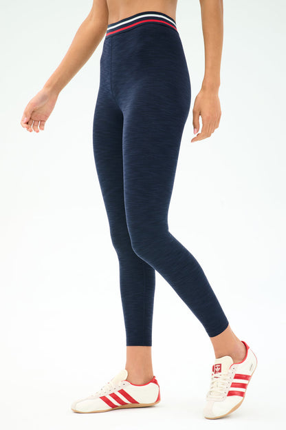 A person wearing SPLITS59 Bailey High Waist Active Rib 7/8 leggings in Heather Indigo with a striped waistband and white sneakers with red stripes, standing against a plain background—ideal for gym workouts.
