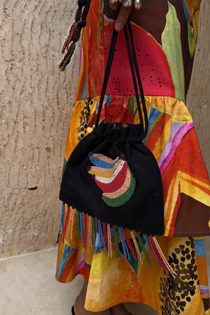 Black Banana Pouch Bag with a beaded motif and multicolour fringe, held over a vibrant fruit-print summer dress.