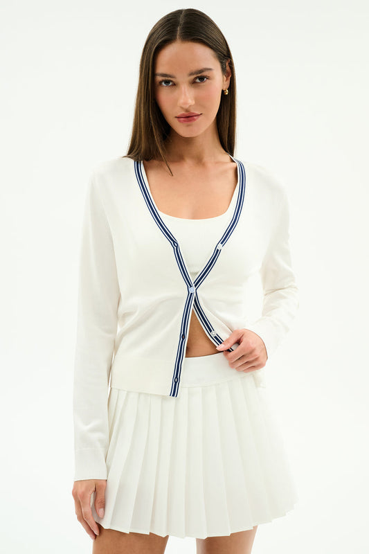 A woman stands against a white background wearing the SPLITS59 Bekah Knit Cardigan in White/Indigo, featuring blue stripes, over a white tank top and a white pleated skirt.