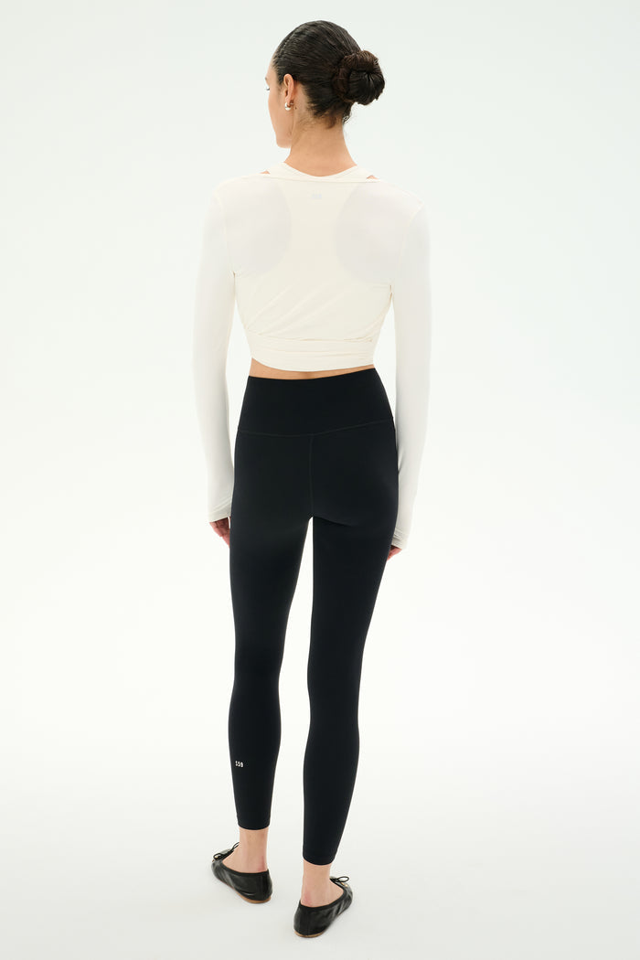A person stands facing away, wearing the SPLITS59 Bella Airweight Lite Wrap in white, paired with high-waisted black nylon-spandex leggings and black ballet flats, against a plain white background.