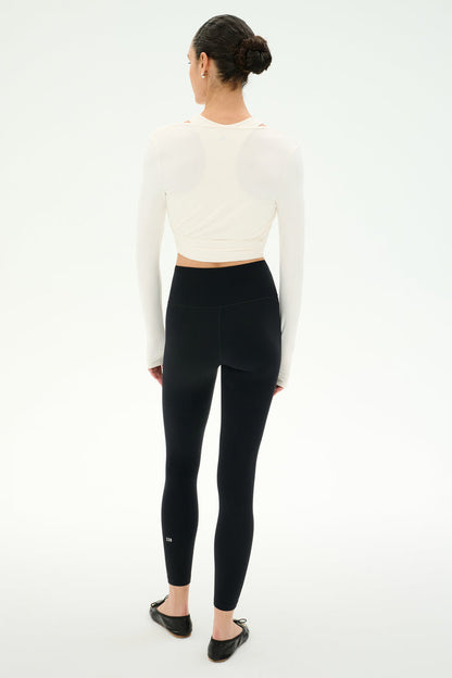 A person stands facing away, wearing the SPLITS59 Bella Airweight Lite Wrap in white, paired with high-waisted black nylon-spandex leggings and black ballet flats, against a plain white background.