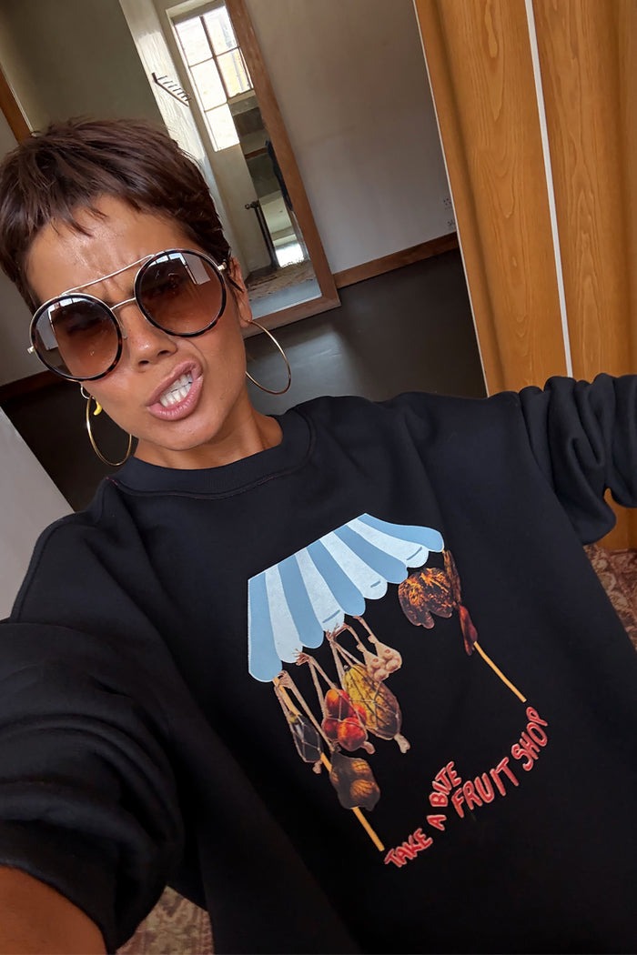 Black Fruit Shop Sweatshirt featuring a blue striped awning and hanging fruit print, worn by a model.