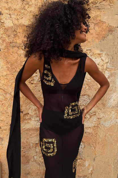 Black & Gold Simona Sequin Dress in sheer mesh with gold motifs and a detachable scarf against a stone wall.