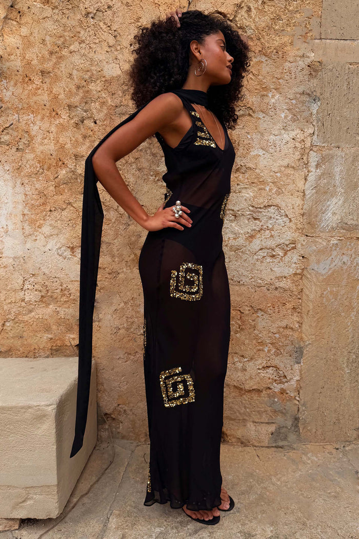 Black & Gold Simona Sequin Maxi Dress in sheer mesh with a trailing scarf, styled on a model against a stone wall.