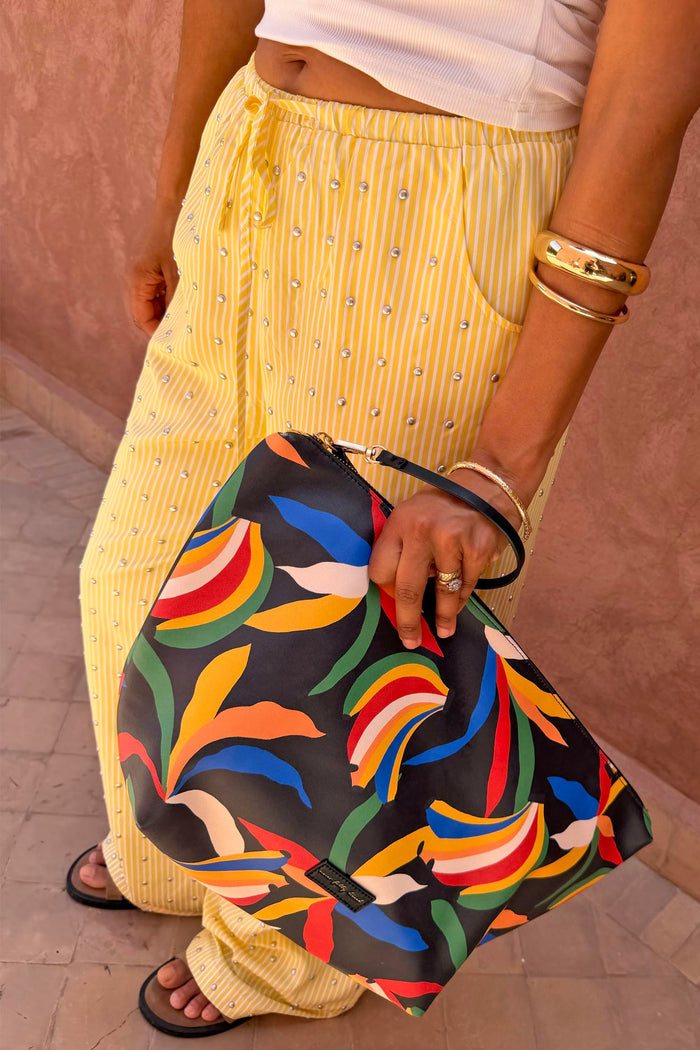 NFD banana print pouch with a black wrist strap, held by a model wearing studded yellow and white striped trousers.