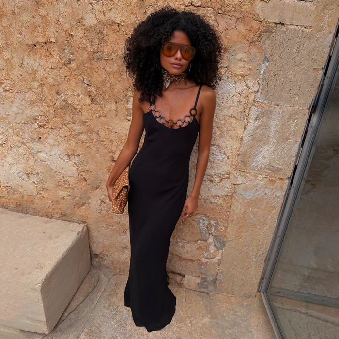 Black Sapna Dress with Ring Detail styled as a maxi dress against a stone wall for a vacation look.