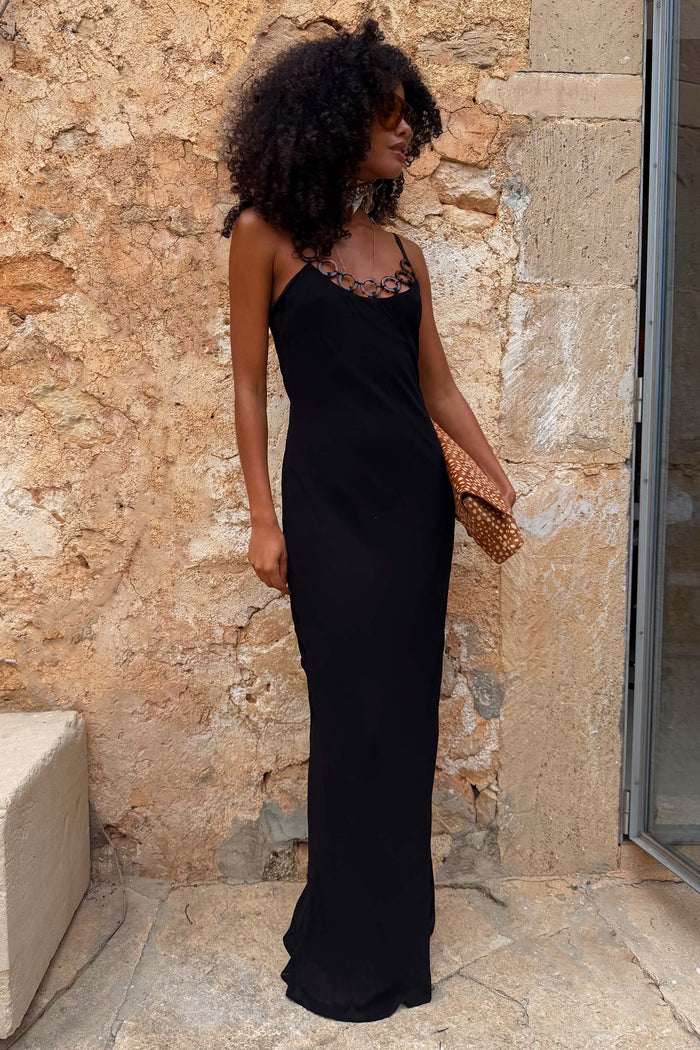 Black Sapna maxi dress with ring detailing at the neckline and a fitted silhouette against a stone wall.