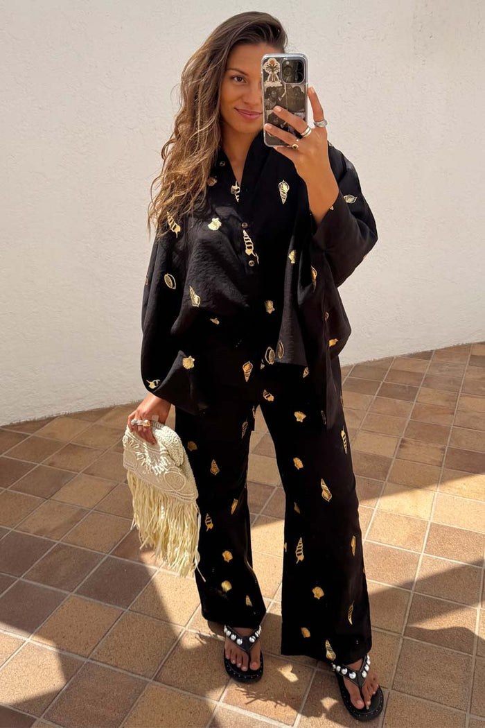 Black Shell Elissa wide-leg trousers with gold shell prints, paired with a matching shirt and a cream fringed bag.