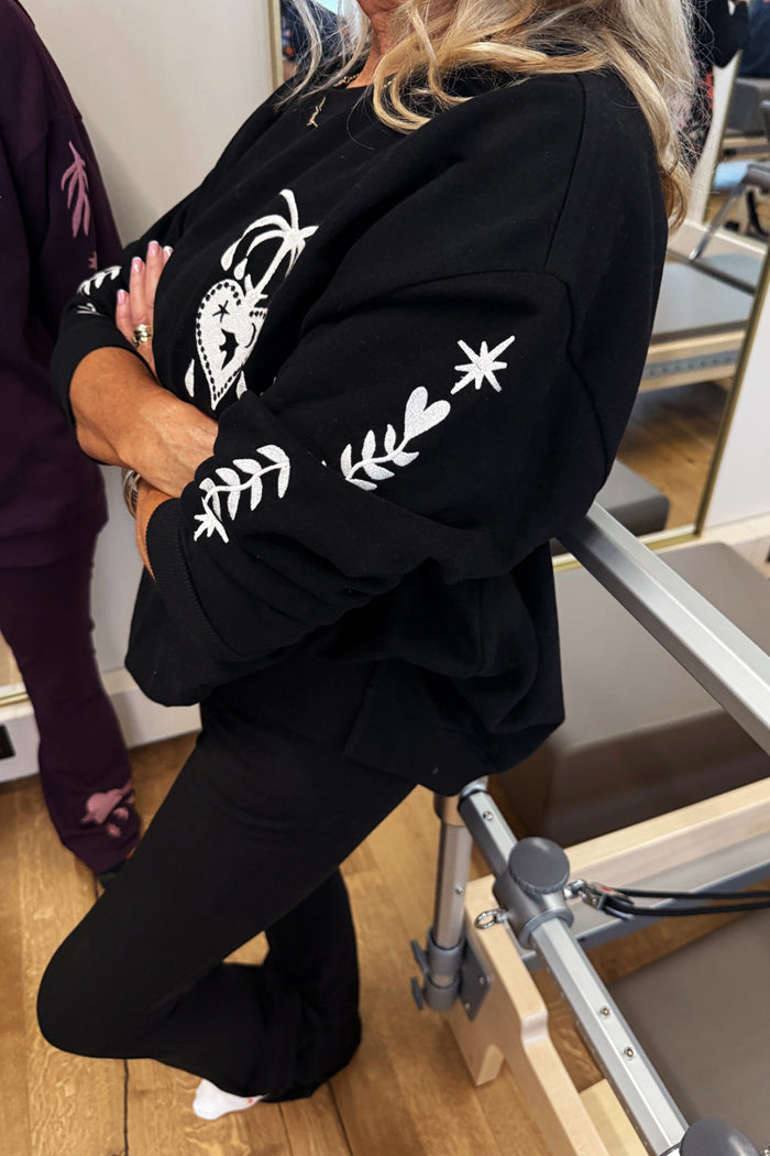 Black sweatshirt with ecru embroidery of heart, star and palm tree motifs on the sleeve, worn at a pilates studio.