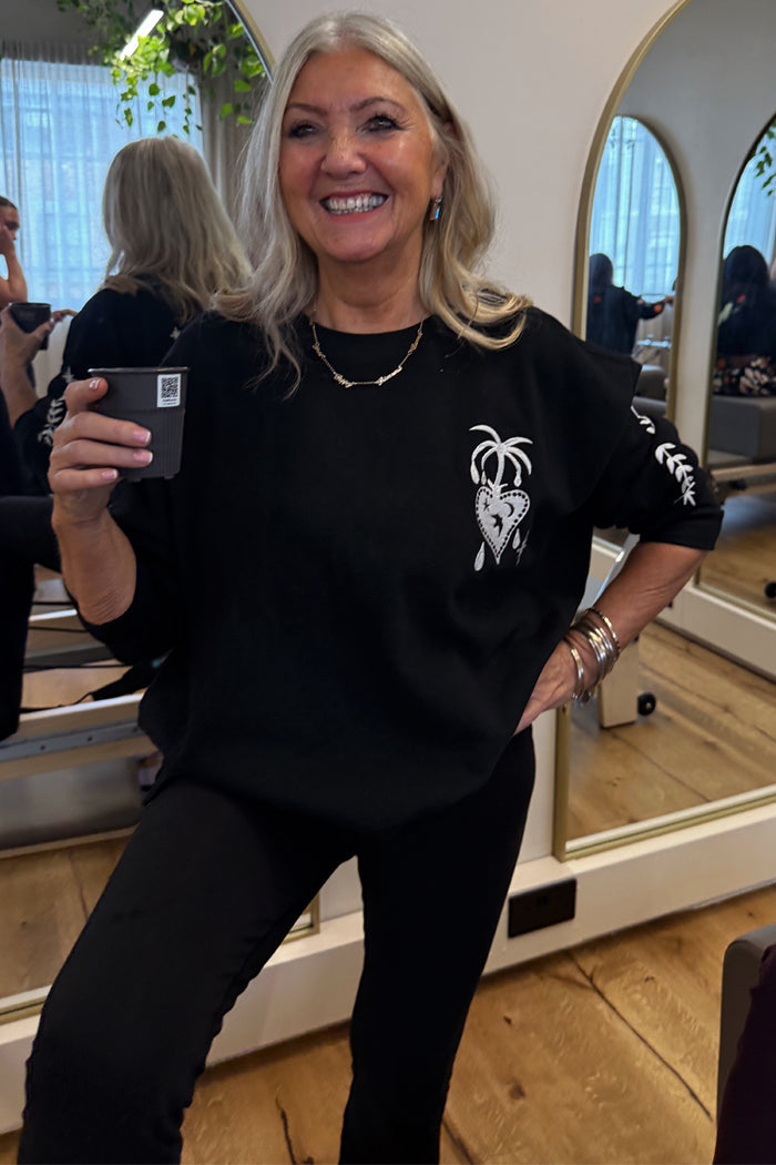 Black Sweatshirt with Ecru Embroidery featuring palm and heart motifs, styled in an oversized fit on a smiling woman.