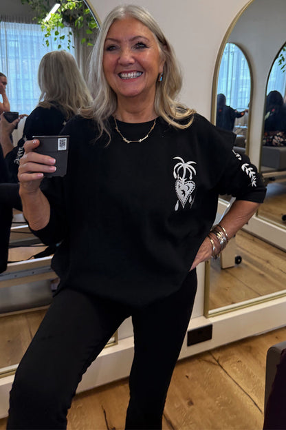 Black Sweatshirt with Ecru Embroidery featuring palm and heart motifs, styled in an oversized fit on a smiling woman.