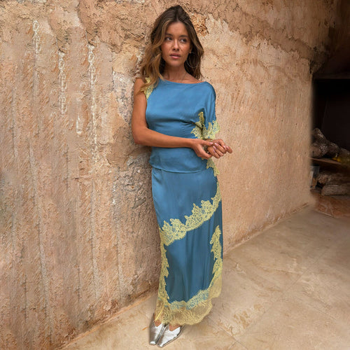 Blue and Lemon Millie Skirt and one-shoulder top co-ord with lace trim, leaning against a stone wall.