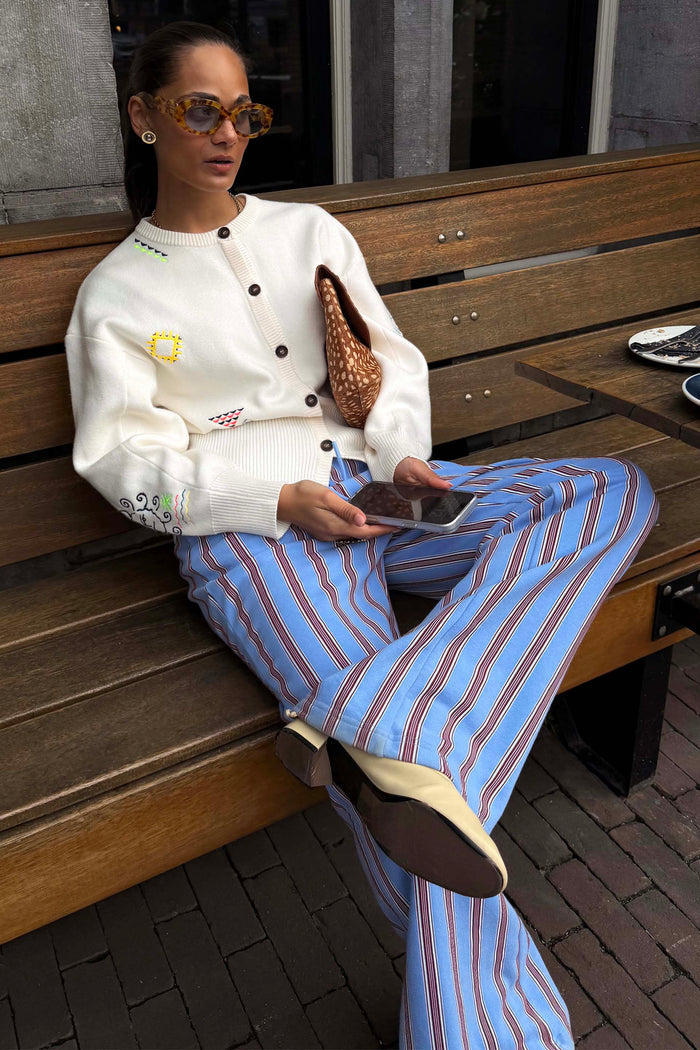 Blue Cotton Stripe Jogger with red pinstripes, styled with a cream embroidered cardigan and tortoise sunglasses while seated on a bench.
