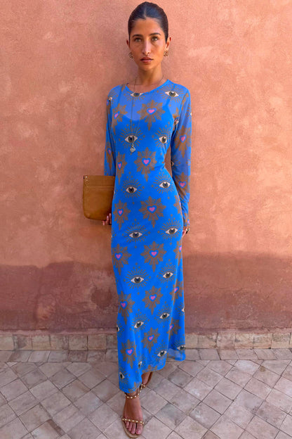 Blue Eye Mesh Maxi Dress with pink heart and eye motifs and long sleeves, worn by a model against a terracotta wall.