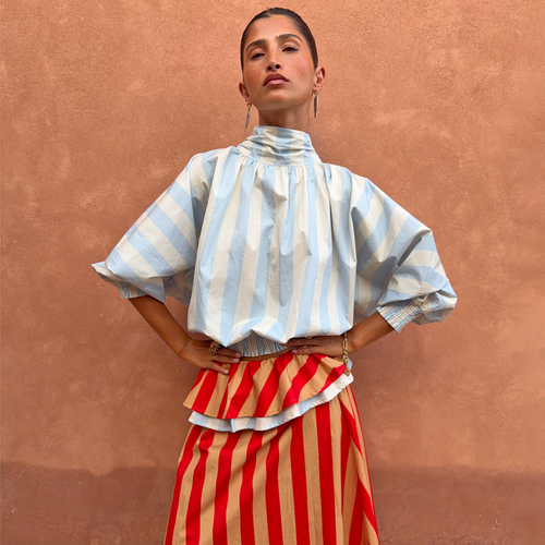 Blue Stripe Batwing Blouse with a high neck and shirred cuffs, worn by a model against a terracotta wall.