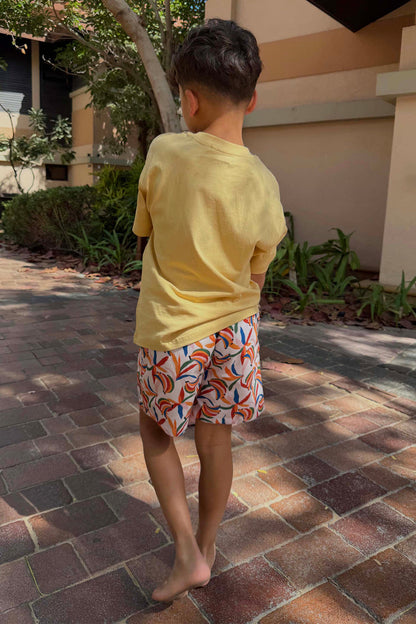 Boy's Banana Print Swim Shorts in cream with multi-coloured fruit, paired with a yellow tee for vacation.