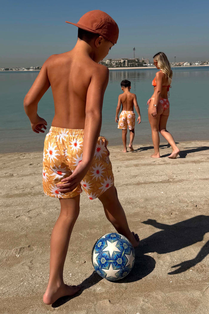 Boy's Yellow Sun Swim Shorts with a white and orange sun print, worn while playing football on a beach vacation.