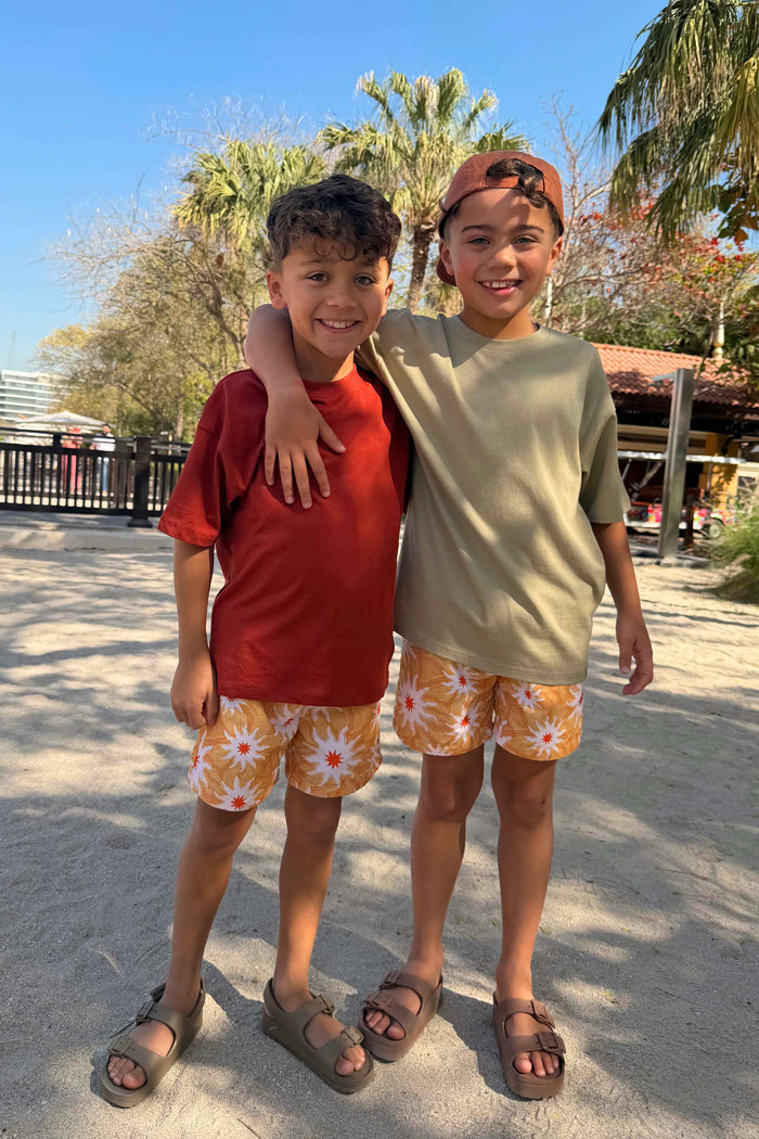 Boy's Yellow Sun Swim Shorts with a sun print and drawstring, worn by two children on a sunny vacation.