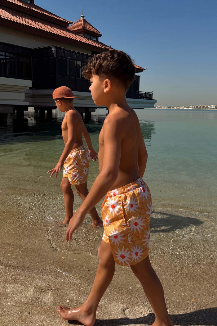 Yellow Sun Swim Shorts with a sun print and drawstring waist, worn by two boys walking in shallow beach water.