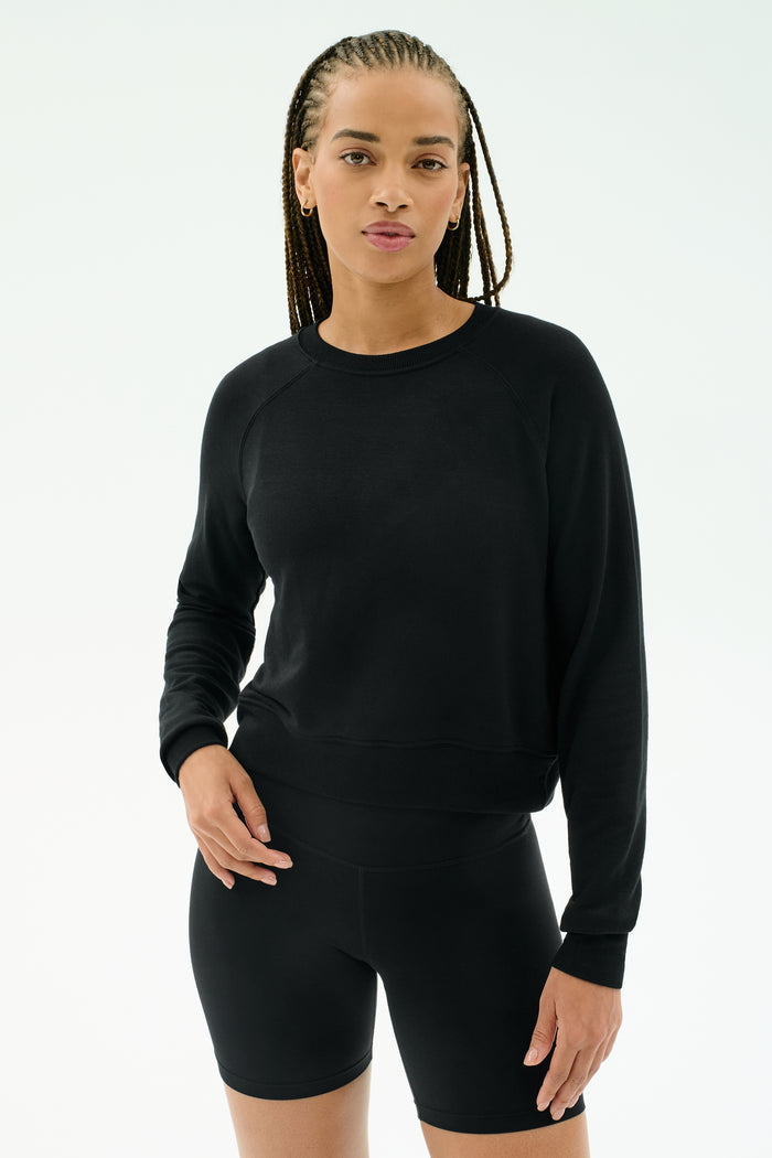A woman wears the SPLITS59 Brooks Fleece Long Sleeve Sweatshirt in black, paired with fitted shorts, standing against a plain white background. Made in Los Angeles.