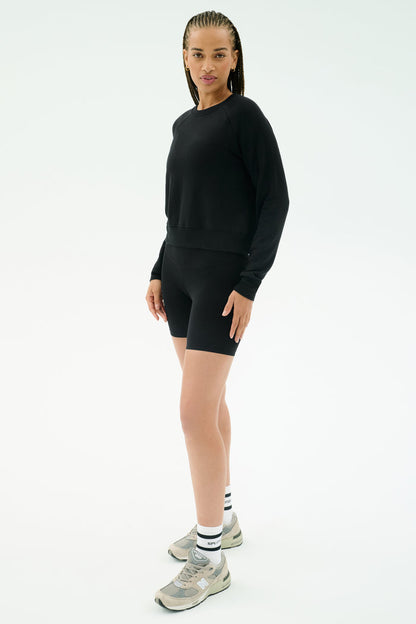 A person wearing the SPLITS59 Brooks Fleece Long Sleeve Sweatshirt in black, black bike shorts, white socks, and gray sneakers stands against a plain white background.