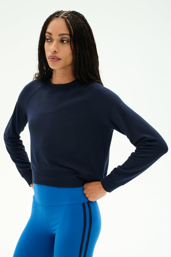 A woman stands with her hands on her hips against a plain light background, wearing the SPLITS59 Brooks Fleece Long Sleeve Sweatshirt in Indigo—a raglan sleeve crewneck pullover. She pairs it with bright blue athletic leggings featuring black stripes for a stylish look made in Los Angeles.