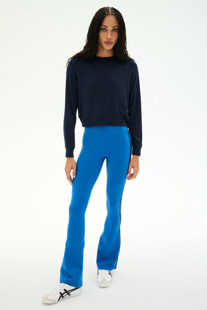 A person stands on a white background wearing the SPLITS59 Brooks Fleece Long Sleeve Sweatshirt in Indigo, paired with bright blue flared pants and white sneakers with black details. This long sleeve pullover is made in Los Angeles.