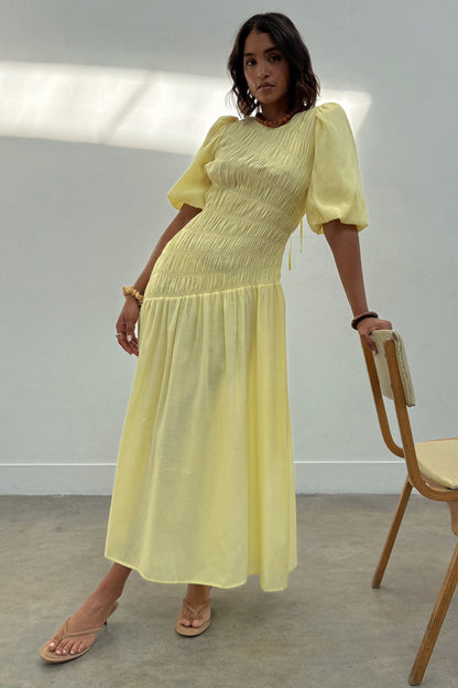 Butter yellow Nara midaxi dress with a shirred bodice, puff sleeves and pleated skirt on a model leaning on a chair.
