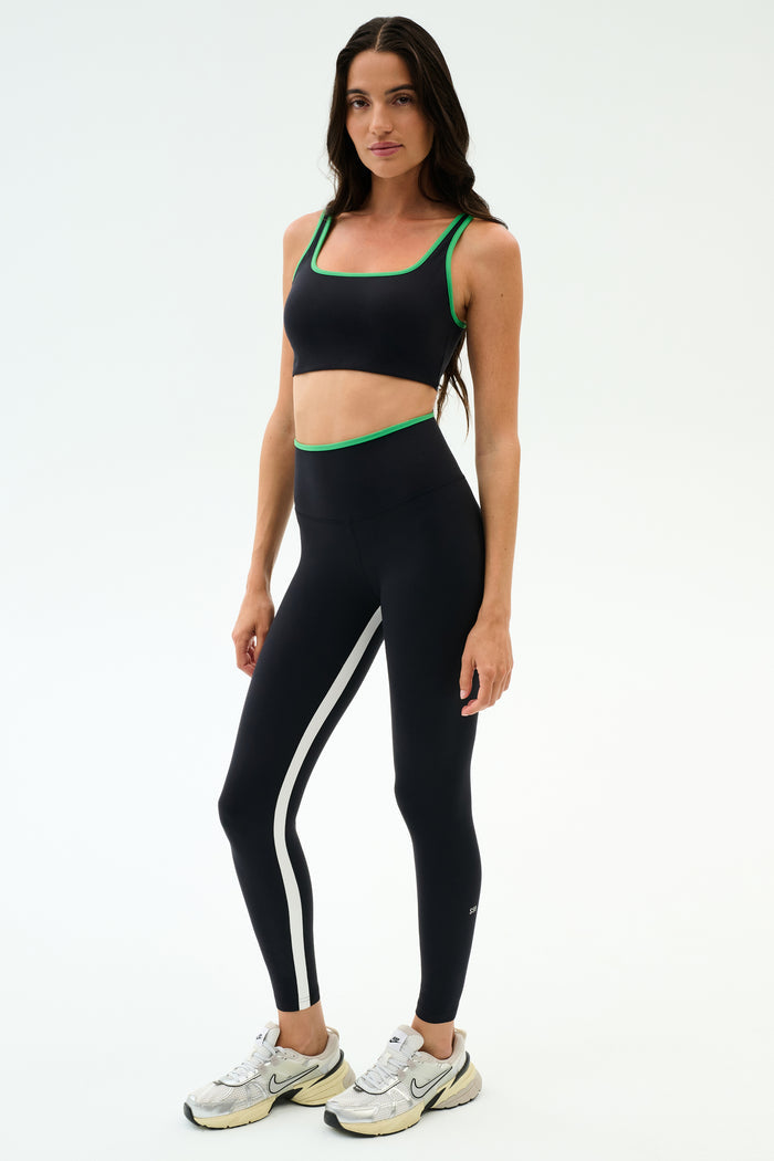 A woman stands against a plain background wearing the SPLITS59 Cait Rigor Bra in Black/Grass, crafted from high-performance Rigor fabric—perfect for high-impact workouts—paired with black leggings and white sneakers.