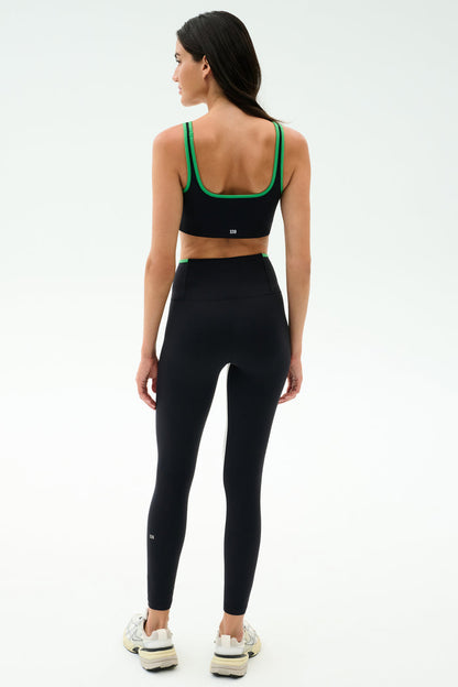 A woman faces away, wearing the SPLITS59 Cait Rigor Bra in Black/Grass, made from high-performance Rigor fabric, paired with high-waisted black leggings and white sneakers against a plain white background.