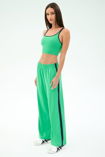 A woman stands against a white background wearing the SPLITS59 Cait Rigor Bralette in Grass/Indigo, matched with wide-leg pants featuring dark side stripes and paired with white sneakers.