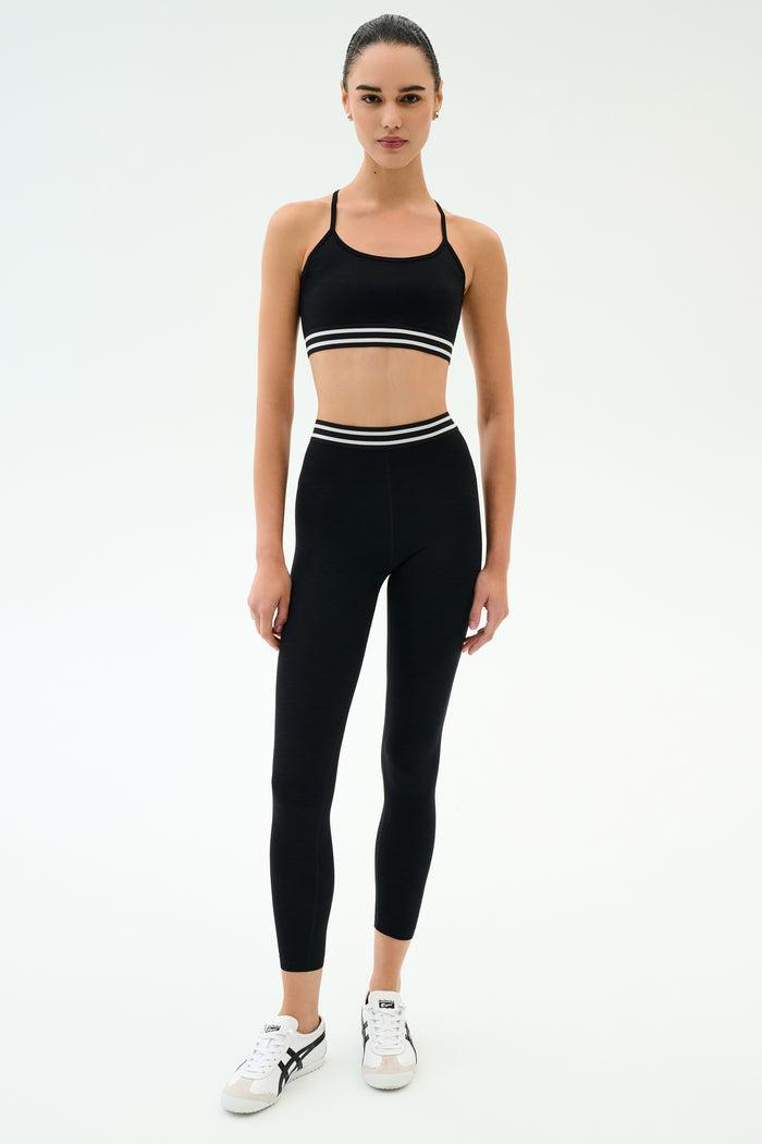 A person wearing the SPLITS59 Cali Active Rib Bra in black and high-waisted black leggings with white stripes, paired with white sneakers, stands against a plain white background.