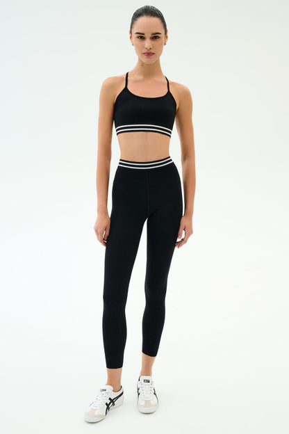 A person wearing the SPLITS59 Cali Active Rib Bra in black and high-waisted black leggings with white stripes, paired with white sneakers, stands against a plain white background.