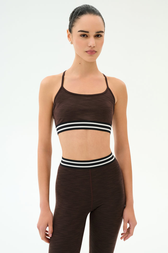 A person stands facing forward, wearing the SPLITS59 Cali Active Rib Bra in Heather Dark Chocolate and matching high-waisted leggings with black and white striped bands.