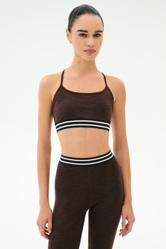 A person stands facing forward, wearing the SPLITS59 Cali Active Rib Bra in Heather Dark Chocolate and matching high-waisted leggings with black and white striped bands.