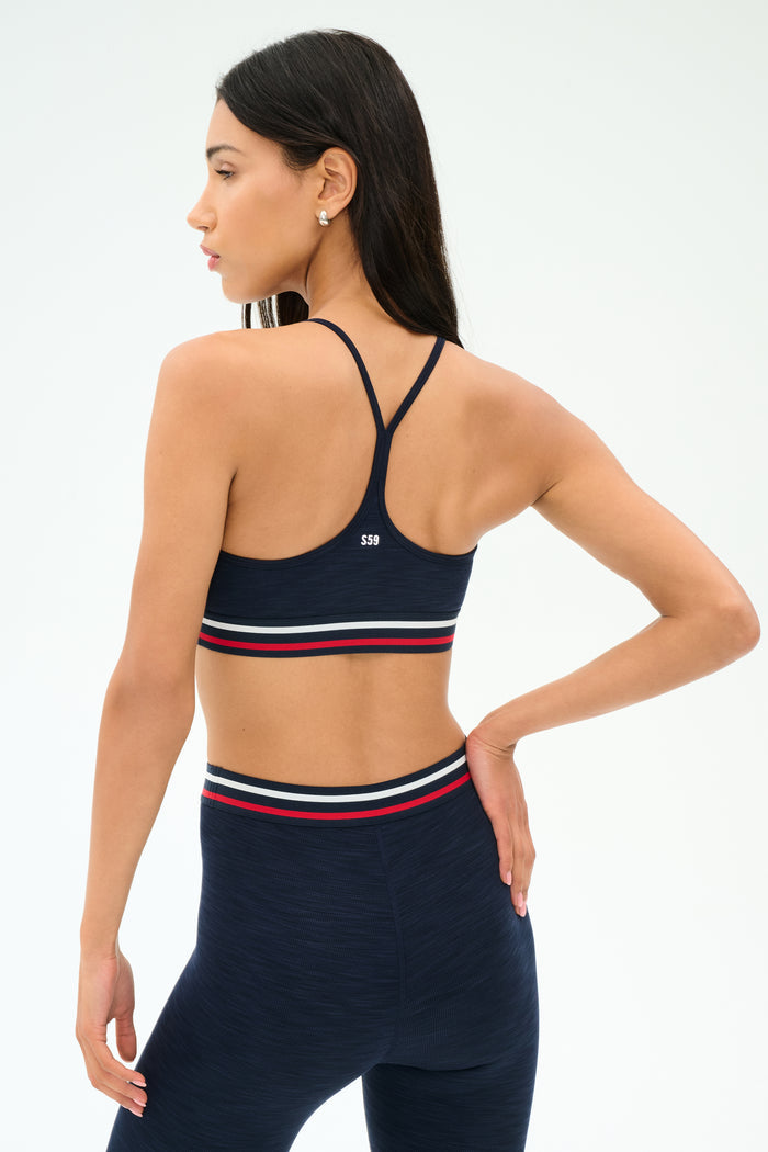 A woman poses from the back against a plain background, wearing the SPLITS59 Cali Active Rib Bra in Heather Indigo and matching high-waisted leggings with red, white, and navy striped bands.