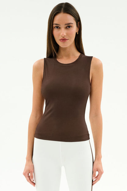 A woman faces the camera in a SPLITS59 Carter Airweight Lite Tank in Dark Chocolate, paired with white pants—ideal Pilates attire—against a plain white background.