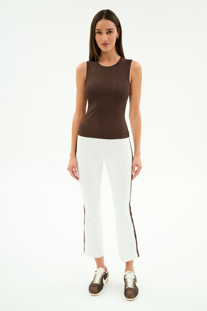 A woman faces forward wearing the SPLITS59 Carter Airweight Lite Tank in Dark Chocolate, paired with white pants featuring brown side stripes and brown sneakers, set against a plain white background.